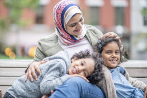 A muslim mother smiles with her children in her lap while sitting on a bench. They are all intertwined and happy to be spending time together. | Une mère musulmane sourit avec ses enfants sur ses genoux, assise sur un banc. Ils sont tous liés et heureux de passer du temps ensemble.