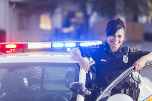 A female police officer getting out of her patrol car. She is smiling, looking at the camera, leaning one arm on the open door of the vehicle. The lights are on. | Une policière sortant de sa voiture de patrouille. Elle sourit, regarde la caméra, appuie un bras sur la portière ouverte du véhicule. Les lumières sont allumées.