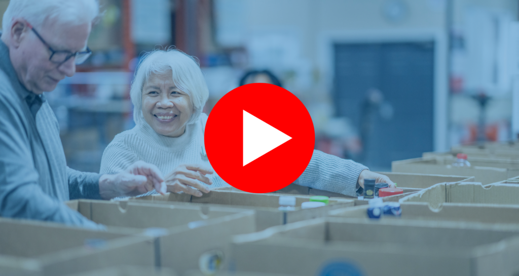A senior gentleman and his wife work together as they pass food items between them and work to sort and pack boxes at a local Food Bank. They are both dressed casually and are smiling at one another as they enjoy their volunteer time together. | Un homme âgé et son épouse travaillent ensemble en se passant des produits alimentaires et en triant et en emballant des cartons dans une banque alimentaire locale. Ils sont tous deux habillés de façon décontractée et se sourient tout en profitant de leur temps de bénévolat ensemble.