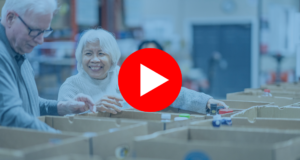 A senior gentleman and his wife work together as they pass food items between them and work to sort and pack boxes at a local Food Bank. They are both dressed casually and are smiling at one another as they enjoy their volunteer time together. | Un homme âgé et son épouse travaillent ensemble en se passant des produits alimentaires et en triant et en emballant des cartons dans une banque alimentaire locale. Ils sont tous deux habillés de façon décontractée et se sourient tout en profitant de leur temps de bénévolat ensemble.