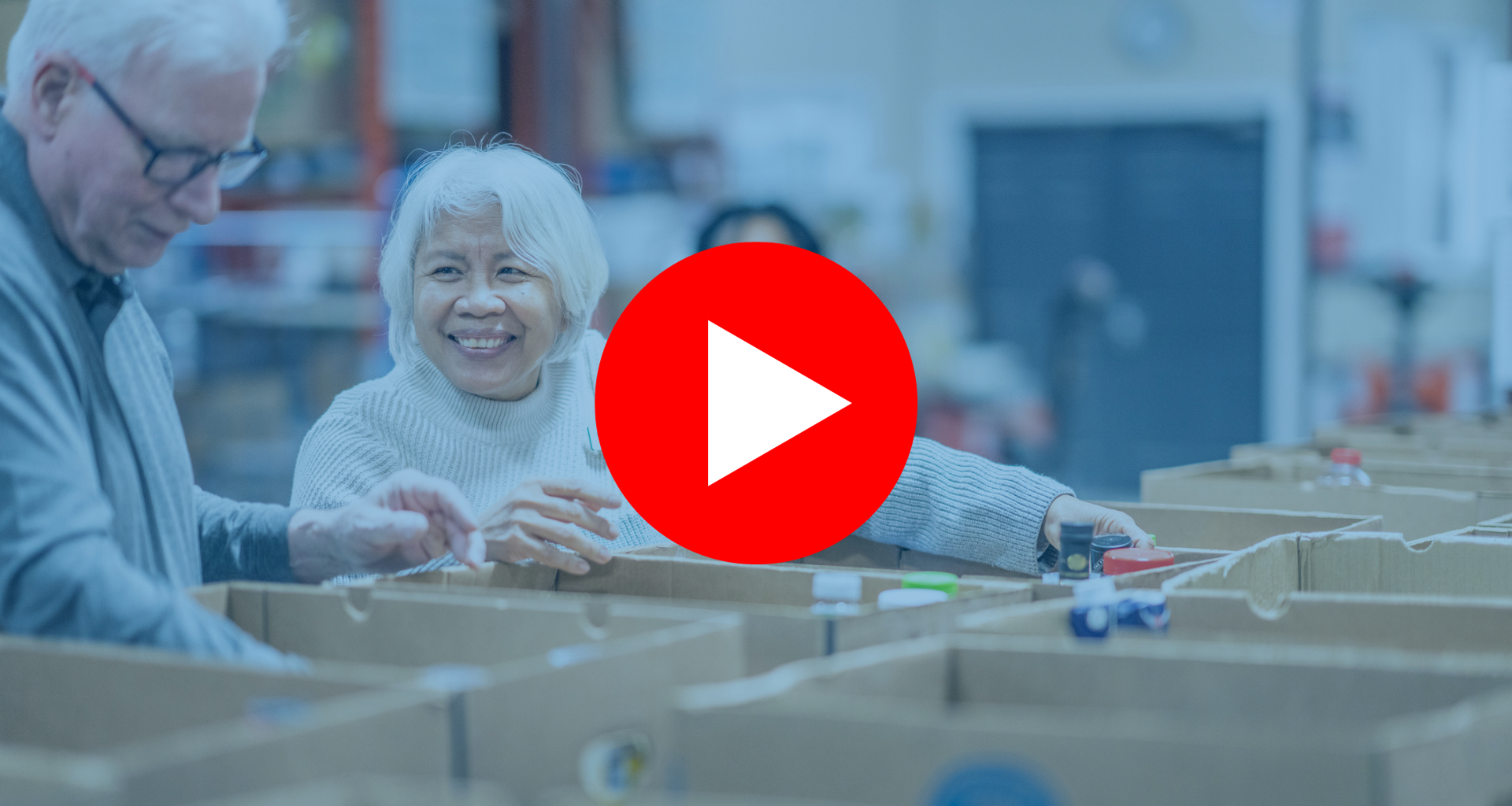 A senior gentleman and his wife work together as they pass food items between them and work to sort and pack boxes at a local Food Bank. They are both dressed casually and are smiling at one another as they enjoy their volunteer time together. | Un homme âgé et son épouse travaillent ensemble en se passant des produits alimentaires et en triant et en emballant des cartons dans une banque alimentaire locale. Ils sont tous deux habillés de façon décontractée et se sourient tout en profitant de leur temps de bénévolat ensemble.