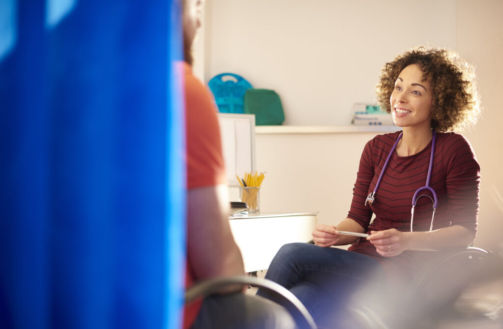 A female doctor sits and chats to a male patient who we see from behind. He is partially obscured by a medical screen. | Une femme médecin est assise et discute avec un patient que nous voyons de dos. Il est partiellement masqué par un écran médical.
