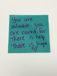 On a teal post-it note, we read the message, "You are valuable. You are cared for. There is help. There is hope." A heart has been drawn beneath the message.