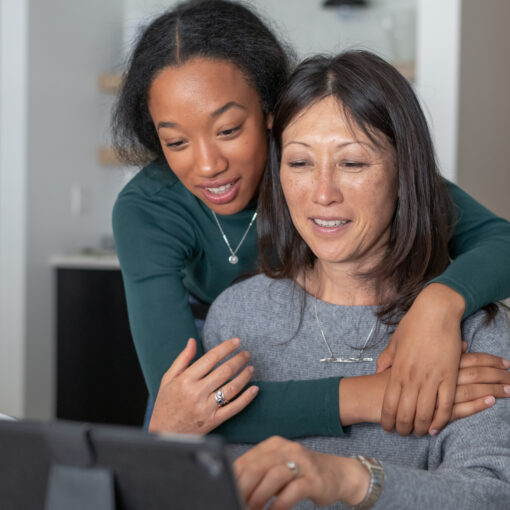 A beautiful mixed race teenager affectionately embraces her mother as they use a tablet computer to visit and stay connected with family and friends while isolating at home during the Covid-19 pandemic. Social distancing, mental health, and social teleconferencing concepts.