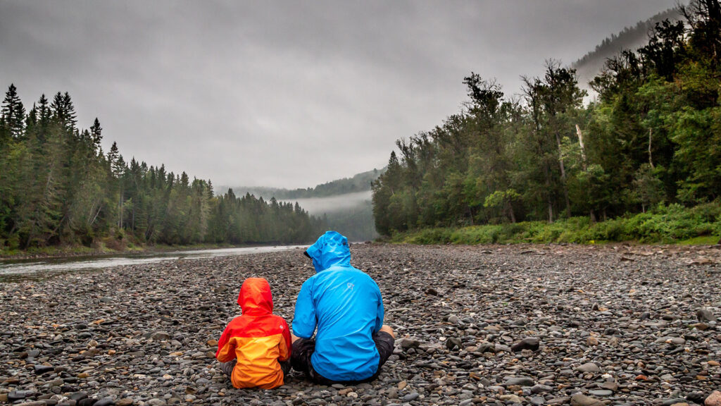 An adult and a child sit on pebbles near the river and look at the mountains
