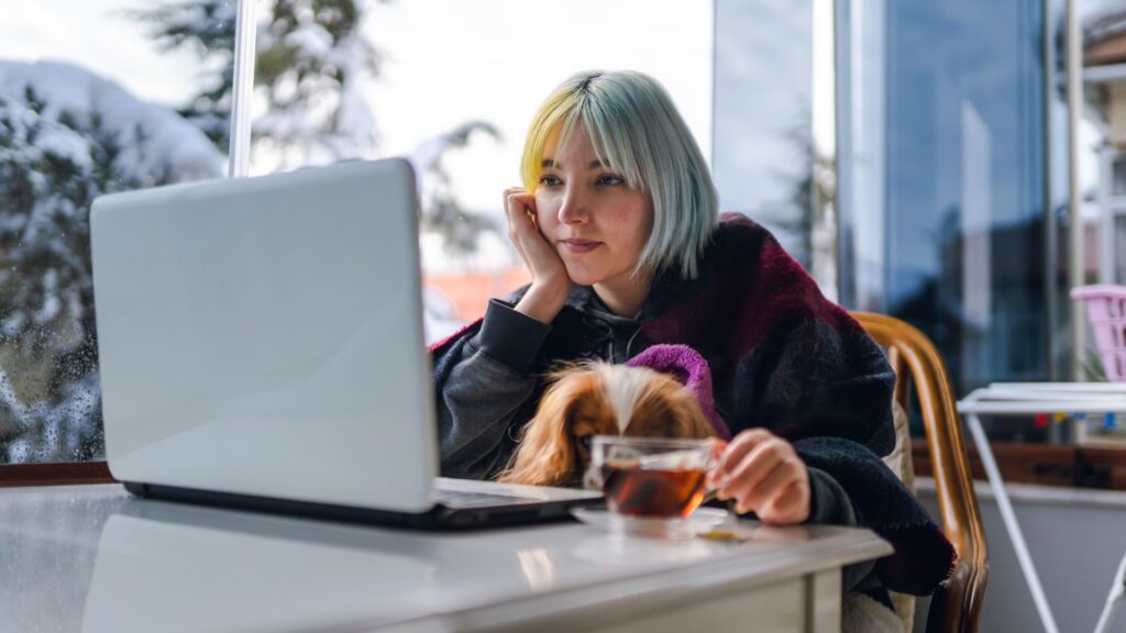 a young woman with a dog on her laps sits in front of the laptop with a cup of tea besides her / une jeune femme avec un chien sur ses genoux est assise devant l'ordinateur portable avec une tasse de thé à côté d'elle