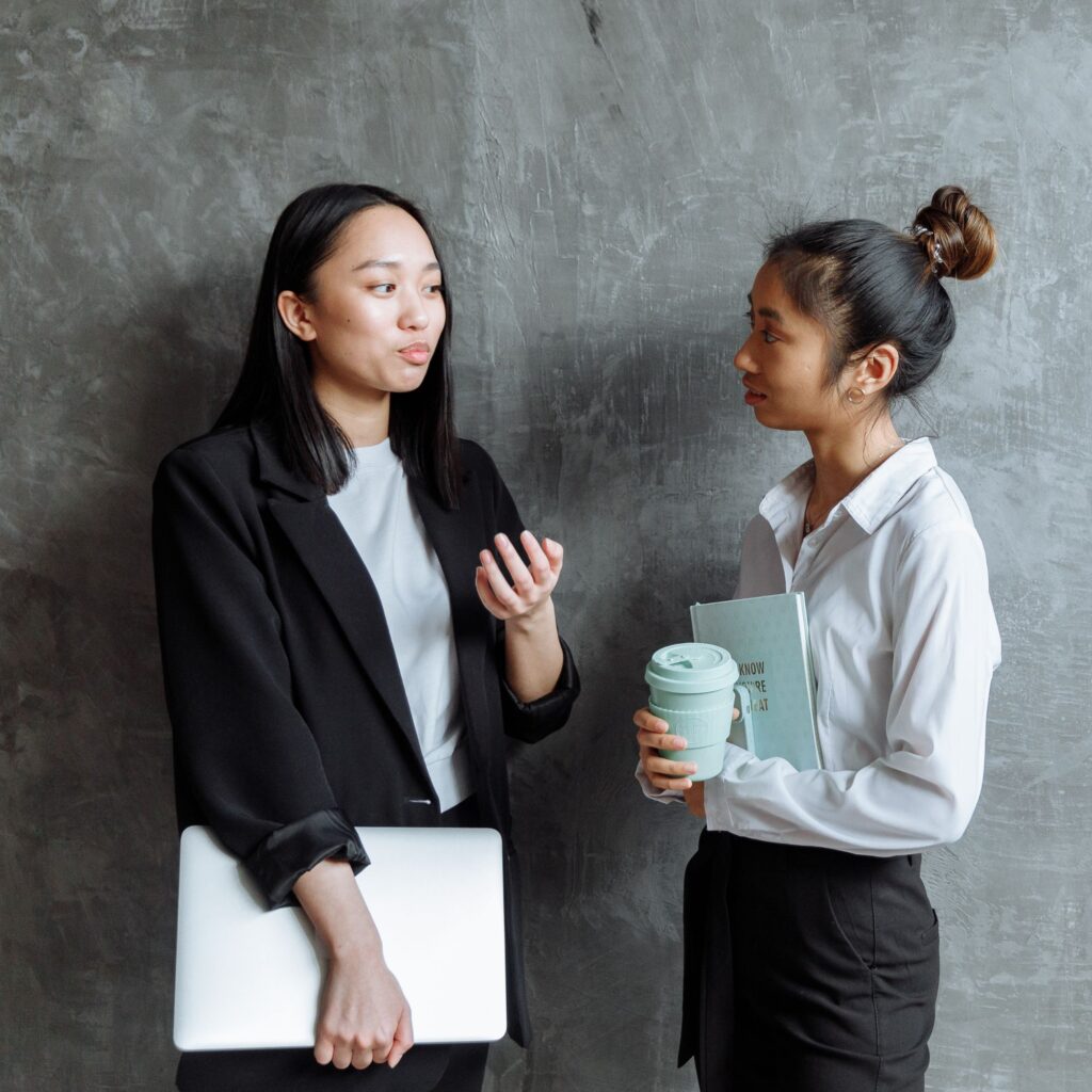 Two young women of Asian decent talking to each other leaning on a grey cement wall