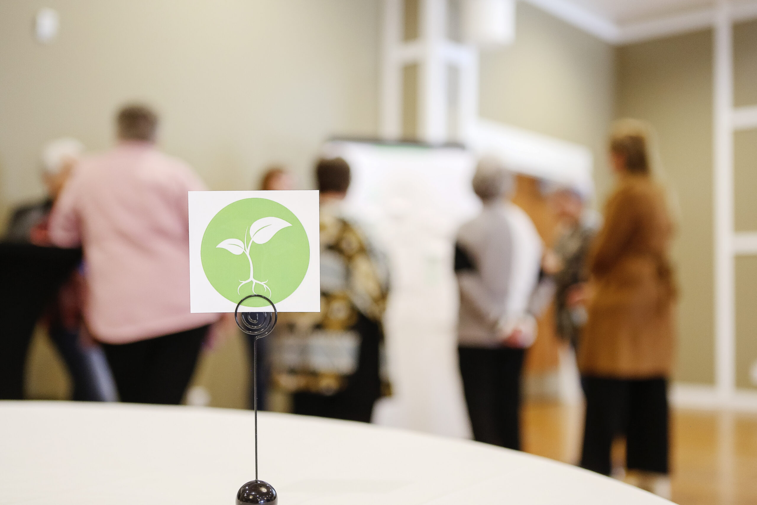 A Roots of Hope table decoration is in the foreground. In the background are blurry people standing in front of an easel. | Une décoration de table Roots of Hope est au premier plan. En arrière-plan, des personnes floues se tiennent devant un chevalet.