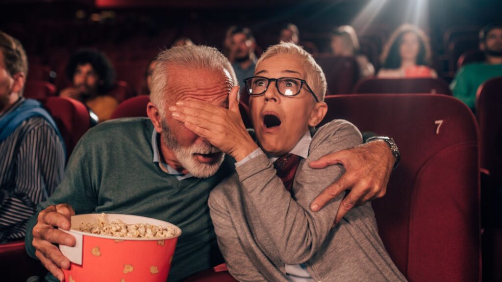 A middle age couple in a movie theater with a popcorn bucket. Women covers man's eyes with her palm