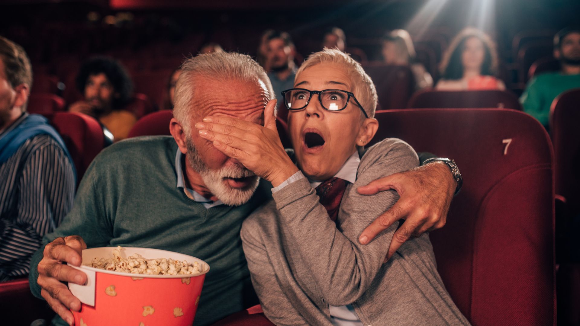 Un couple d'âge moyen dans une salle de cinéma avec un seau de pop-corn. La femme couvre les yeux de l'homme avec sa paume
