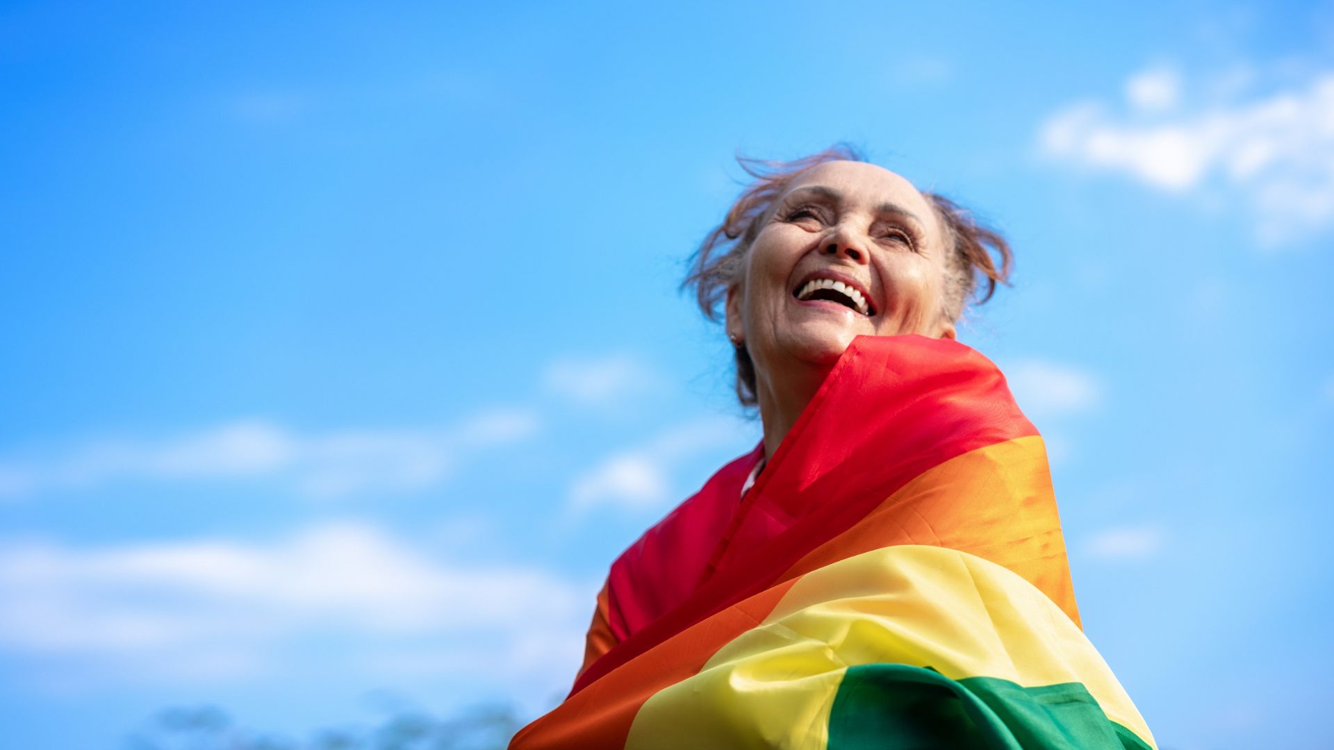 An older woman wrapped in a rainbow-colored flag smiles