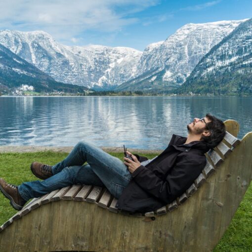 A man is sitting on a wooden chair in front of a lake.