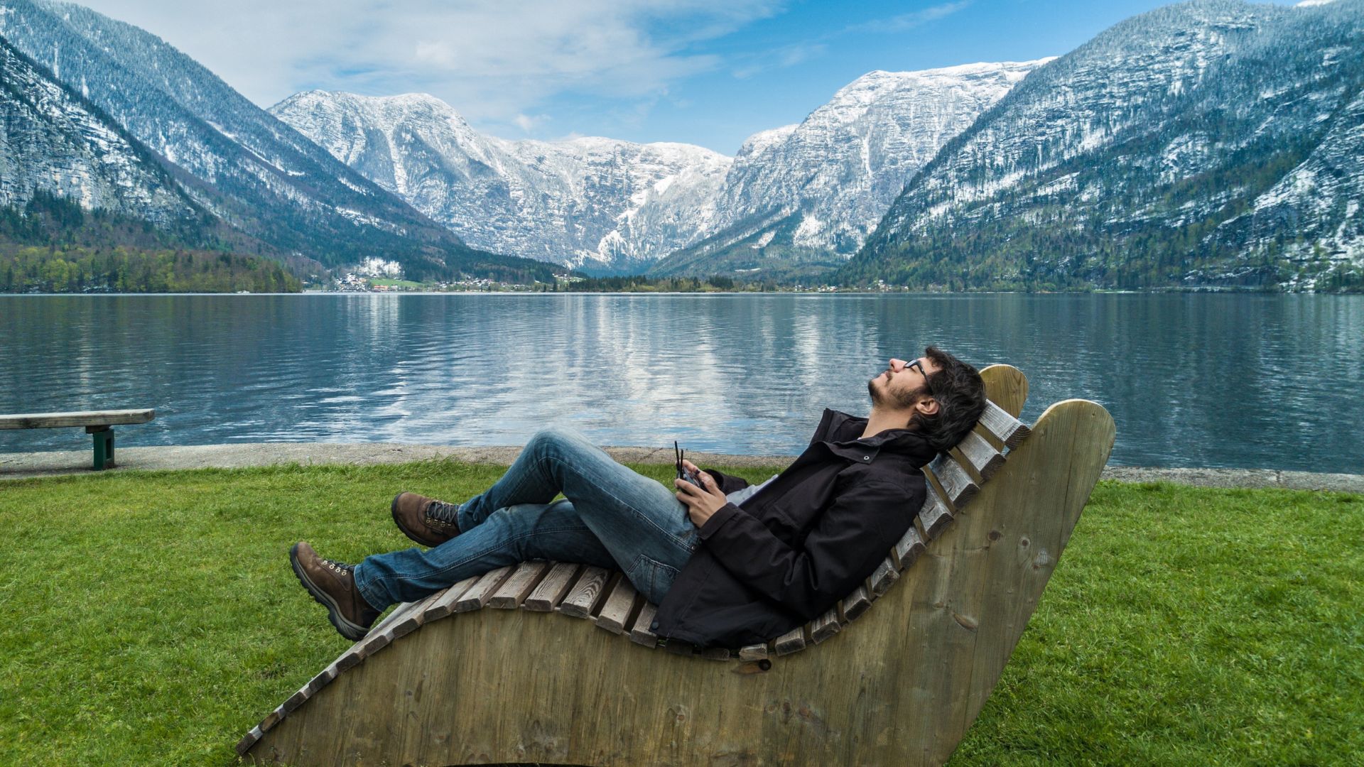 A man is sitting on a wooden chair in front of a lake.