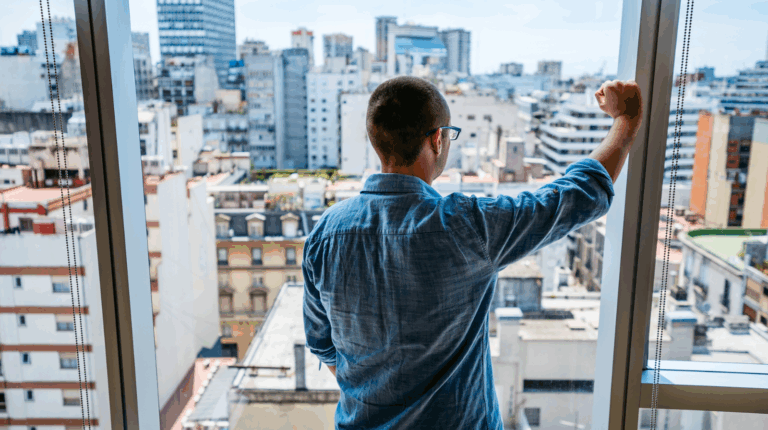 A man is looking out of an apartment window, staring at the city.