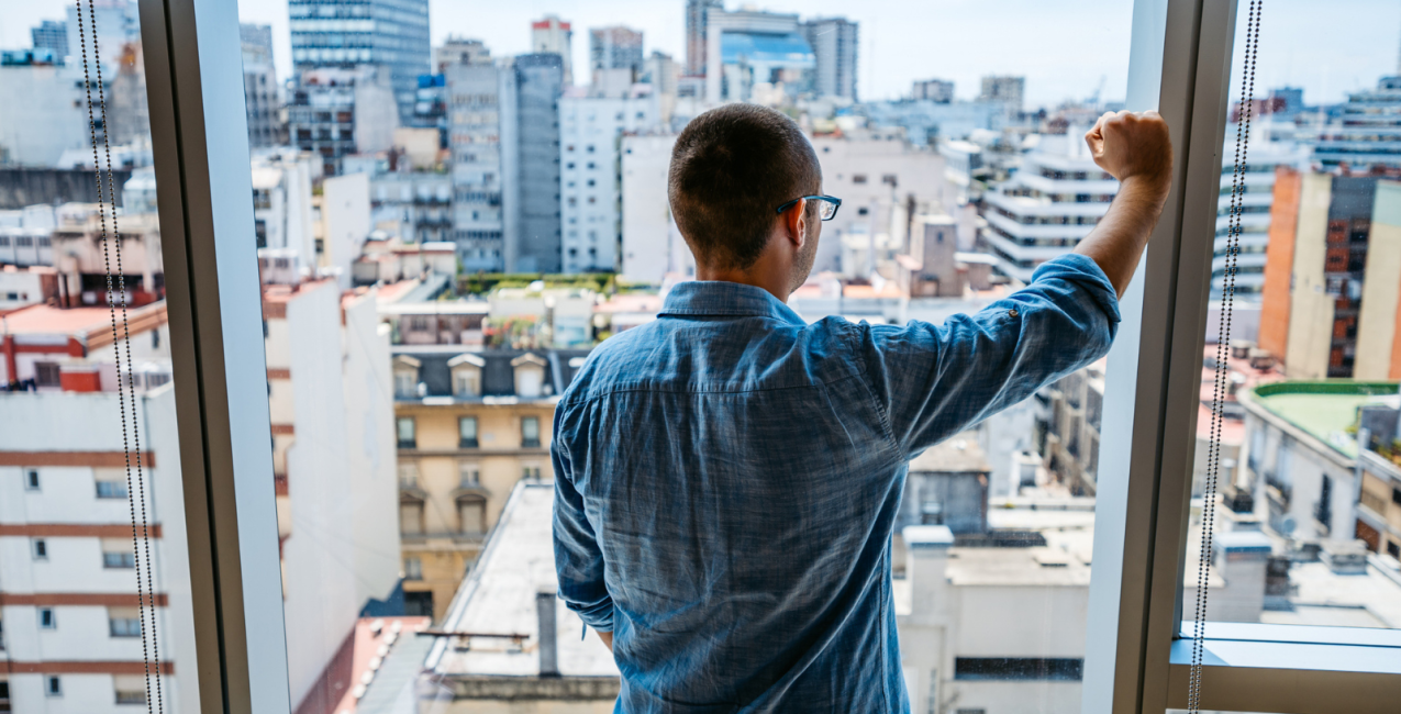 Main Image A man is looking out of an apartment window, staring at the city.